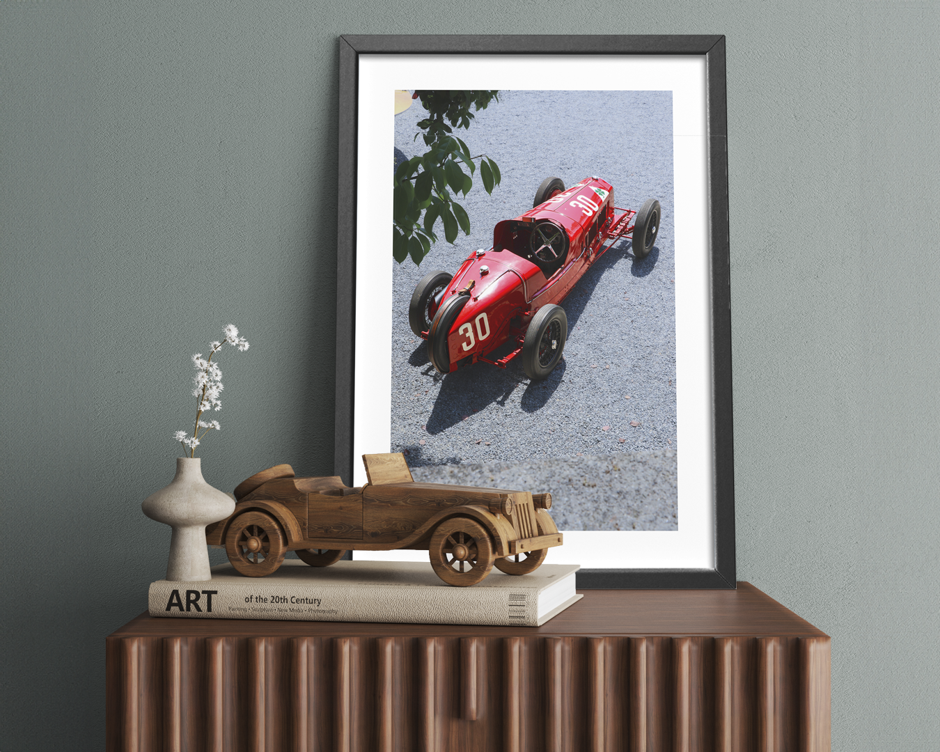 A framed photograph of a vintage red Alfa Romeo 159 race car with the number 30, sitting on a table next to a wooden model car and a book titled "ART of the 20th Century".